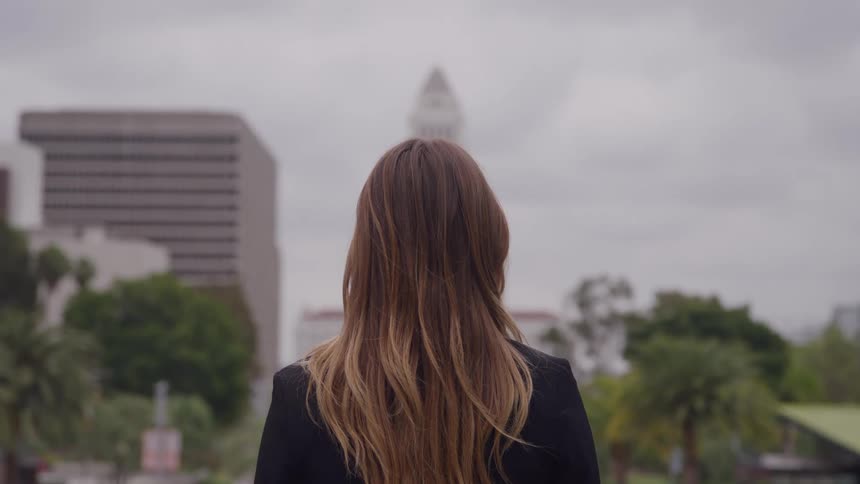 The back of a woman with long brown and blonde hair wearing a black blazer is shown standing outdoors as she looks across a city square toward the downtown skyline on a cloudy day