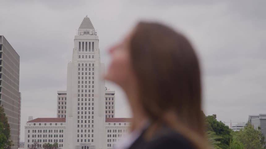 The video shows a blurred closeup of a womans face and shoulder in the foreground looking up towards the skyin the background out of focus is a distinct large white governmentstyle building likely a city hall with a tall central tower visib