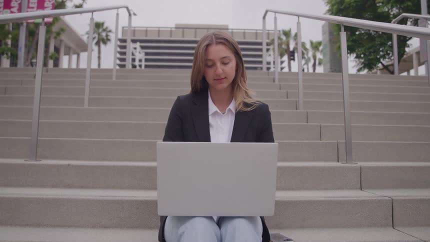 Young woman sitting down on a staircase using a laptop on an urban area outdoors during day time