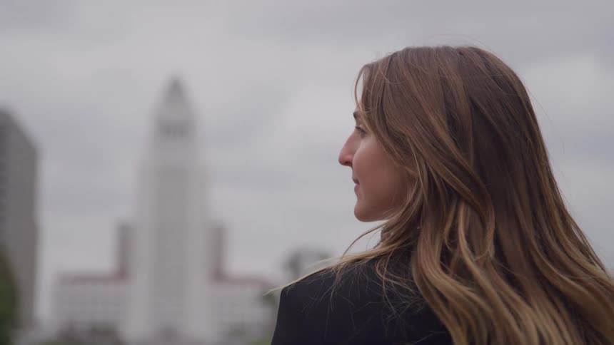 A medium closeup shot focuses on the side profile of a woman with long highlighted hair who looks intently toward a tall blurry landmark building in the background under a uniformly gray overcast sky