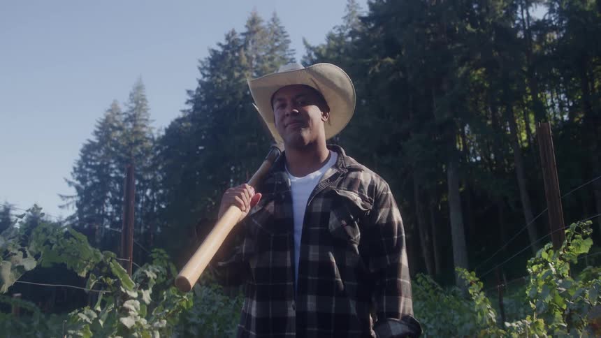 Man in a field holding a tool resting on his shoulder while wearing a widebrimmed hat