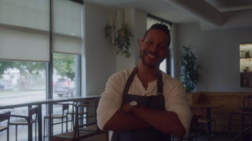 Young adult man wearing a white shirt and a blue apron crossing his arms and smiling at an empty restaurant indoors during day time