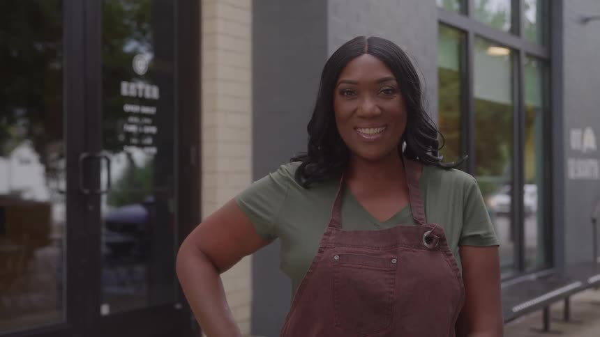 A smiling woman with dark hair wearing a green shirt and a maroon apron stands outside a storefront in daylight