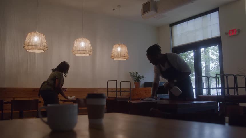 A man and a woman who are coworkers clean and wipe down wooden tables inside a dimly lit cafe with wicker pendant lights and coffee cups visible in the foreground