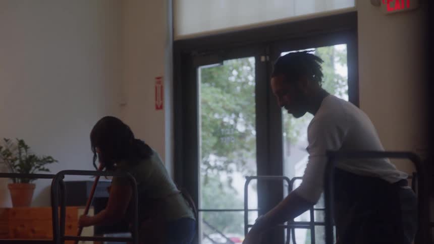 A man and a woman are performing cleaning duties inside a cafe with the man focused on stacking chairs onto a rolling cart and the woman using a broom to sweep the floor as they work collaboratively indoors next to the doorway