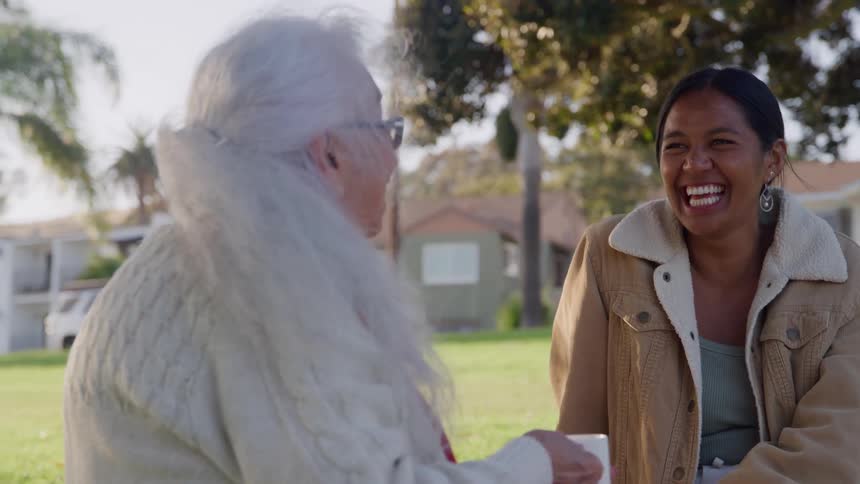 A young woman wearing a tan jacket laughs happily while sitting on the grass outdoors and chatting with an elderly woman with long white hair