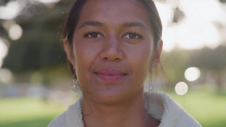 A young woman wearing a creamcolored fleece jacket and earrings looking directly at the camera with a gentle content smile all against a softly focused outdoor background