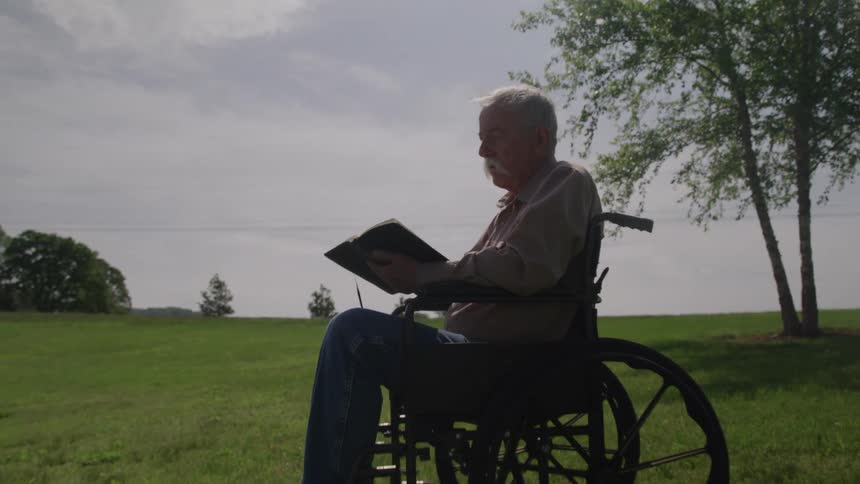 An elderly man reading a book while sitting in a wheelchair outdoors by a pond during daytime