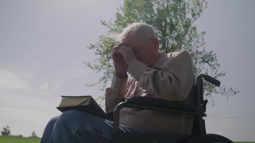 The man sitting in a wheelchair is praying with the bible on his lap