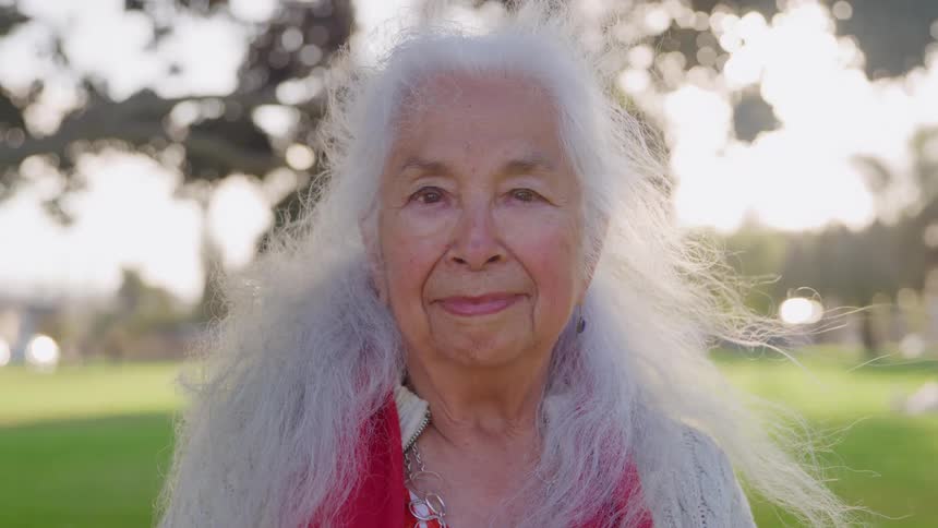 A smiling elderly woman with very long white hair and a red top stands in a sunny park with sun flare peeking through the trees