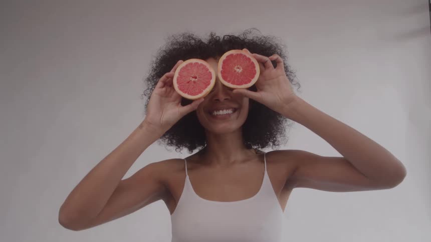 A black woman with curly hair wearing a white tank top smiles broadly and holds two grapefruit halves over her eyes looking forward against a bright neutral background indicating playfulness