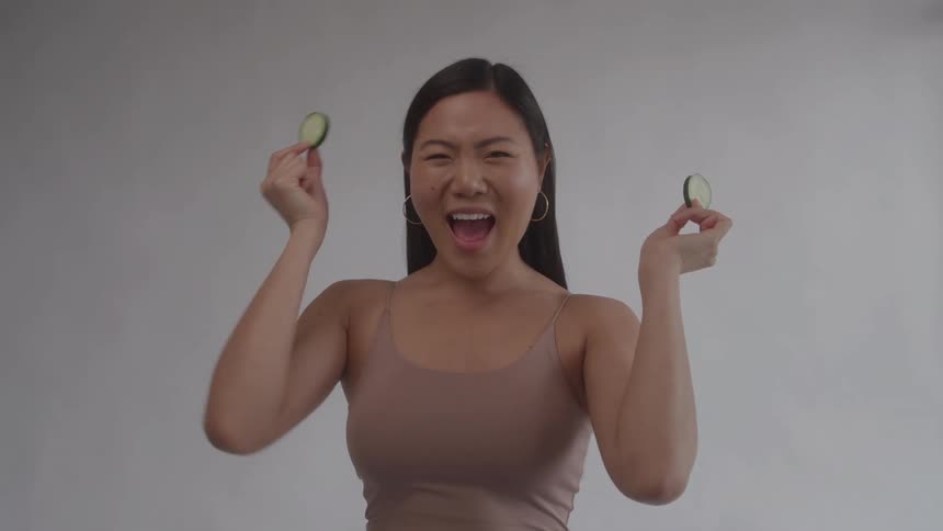 A medium shot of a cheerful woman in a beige tank top holding two slices of fresh cucumber over her eyes and laughing happily against a gray background