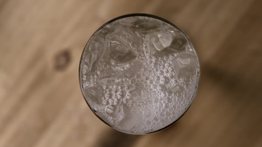 From a direct overhead perspective sparkling bubbles rise rapidly around floating ice cubes in a clear glass fizzing with active carbonation against a soft wooden background