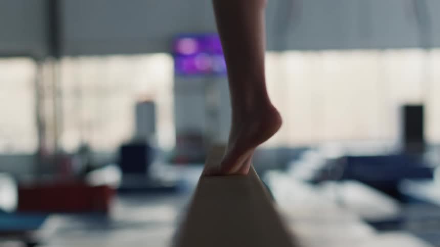 Lower legs and feet of a white woman twisting on one foot over a balance beam in a gym indoors during day time