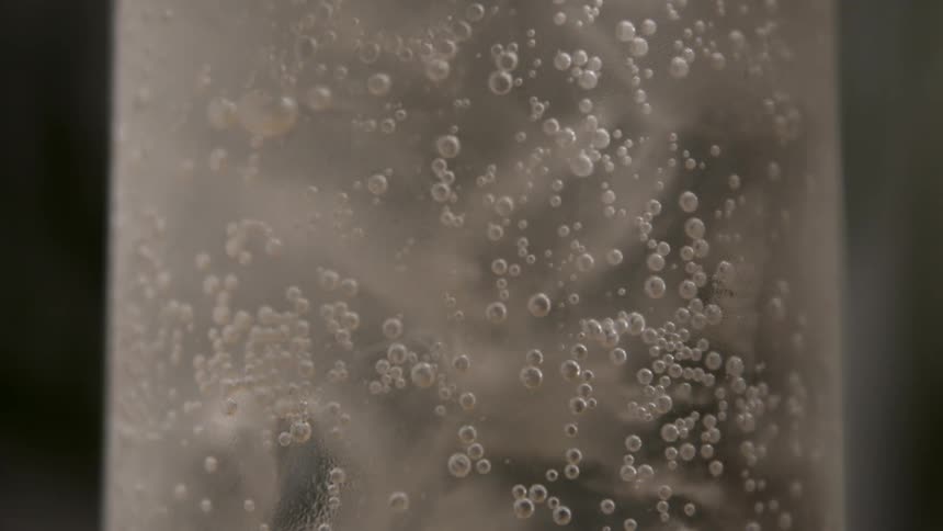 This is a macro closeup shot of the inside of a glass filled with a clear carbonated beverage and ice tiny effervescent bubbles cling to the sides of the glass shimmering against the ice
