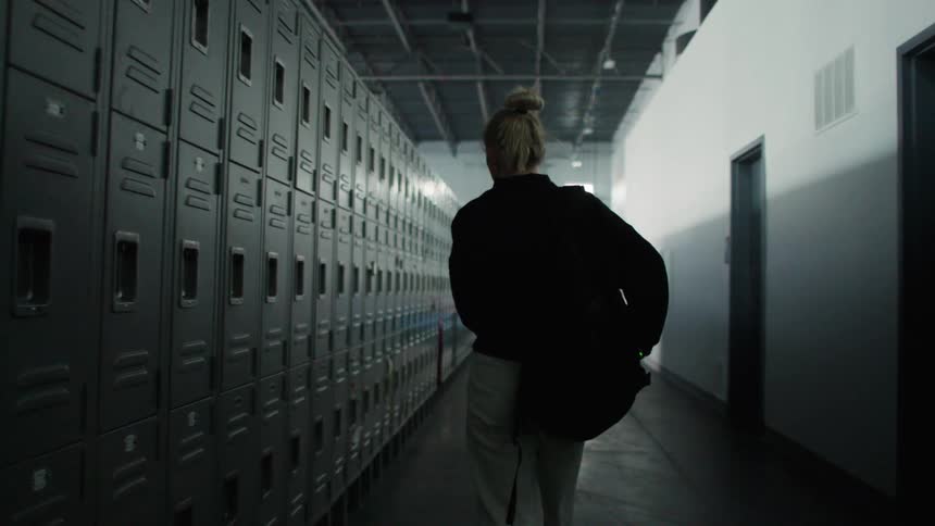 This is a tracking shot from behind a person with their blonde hair pulled up wearing a dark jacket and light pants walking down a narrow hallwaythe hallway is lined with dark grey lockers on one side and a white wall with dark doorways on the other