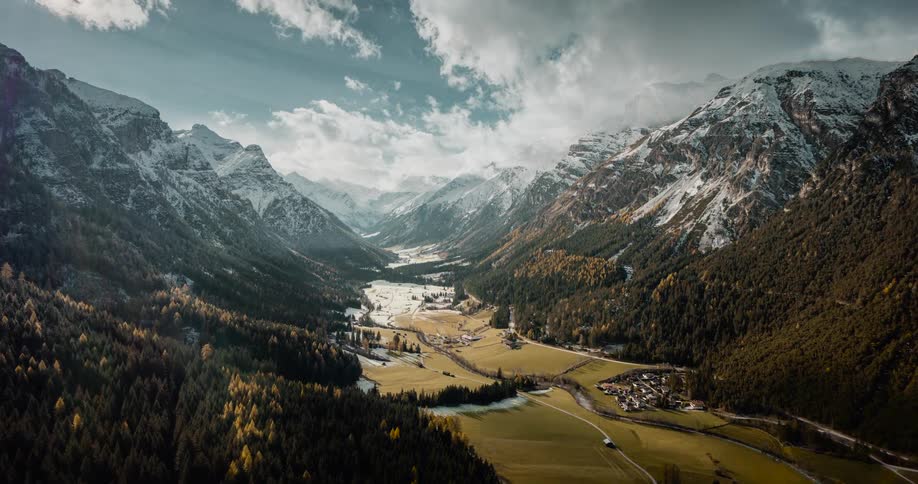 A vast alpine valley is framed by towering mountains with snowdusted peaks leading down to a mosaic of vibrant green meadows and dense evergreen forests