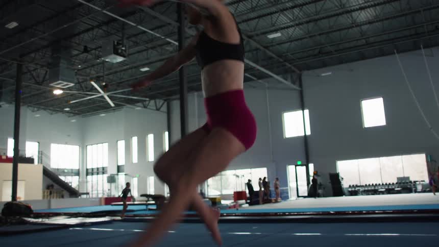 A blonde female athlete with her hair pulled back wearing a black sports bra and red athletic shorts performs a jump followed by a turn on a large blue floor mat in a expansive gymnasium with high ceilings and other girls training in the background
