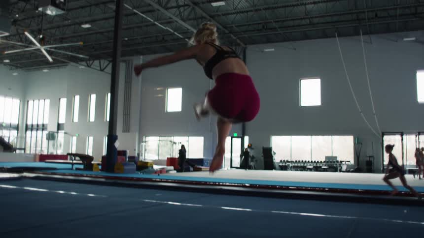 A young female athlete in a black sports bra and red shorts performs powerful split jumps and dynamic movements on a blue floor mat in a brightly lit gymnastics training facility where other athletes and coaches are visible in the background