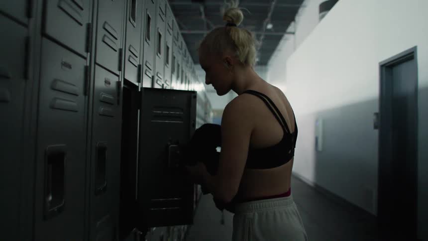 A young athletic woman with her hair in a bun takes off a dark jacket and puts her belongings into an open gray locker in a brightly lit gymnasium locker room