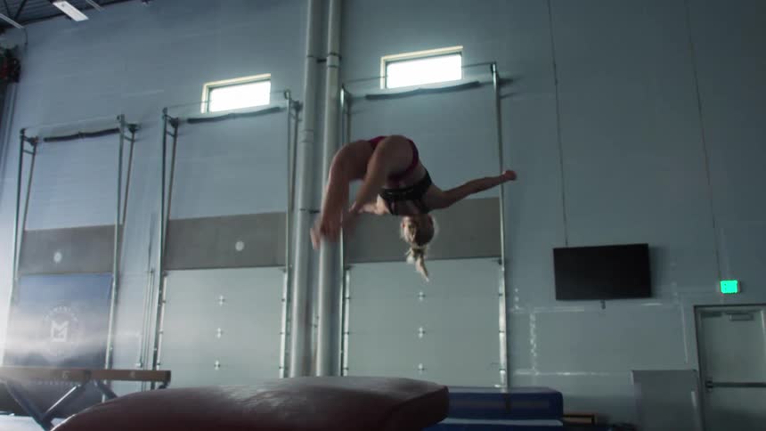 A focused female gymnast wearing a black sports bra and red shorts executes an acrobatic flip or handspring from the top of a vaulting horse and lands on a blue mat performing the maneuver within a highceilinged indoor gymnastics training facility