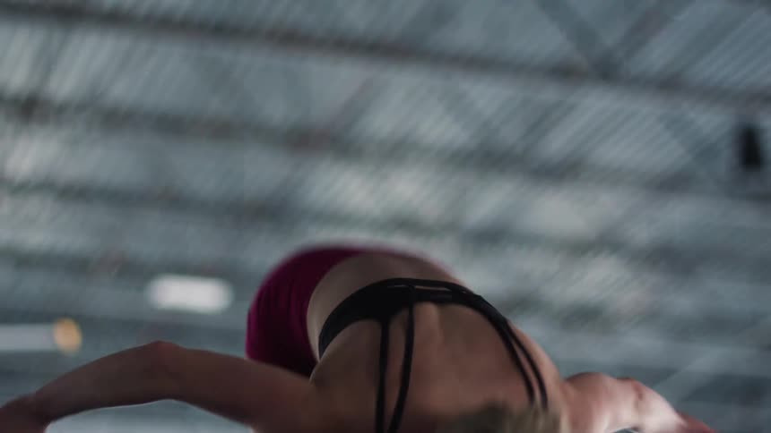 A low angle shot of a focused young athletic woman in a black sports bra and red shorts performing an energetic jump with her arms raised in a gym with a corrugated metal ceiling