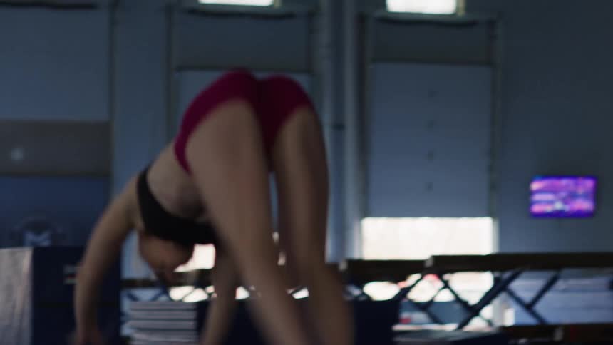 A female gymnast runs and performs a powerful tumbling pass starting with a run a high jump and transitioning into a complex series of flips and turns she is barefoot wearing a black sports bra and maroon shorts and has her hair tied up