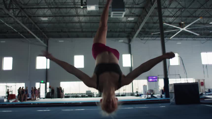 A female gymnast in a maroon leotard is shown doing a flip in a large brightly lit indoor gymnasium with high ceilings and other people in the distance