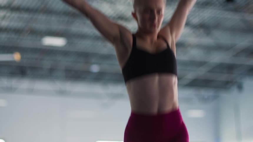 An athletic woman wearing a black sports bra and red leggings raises her arms overhead while exercising in a gym with a blurred industrial background
