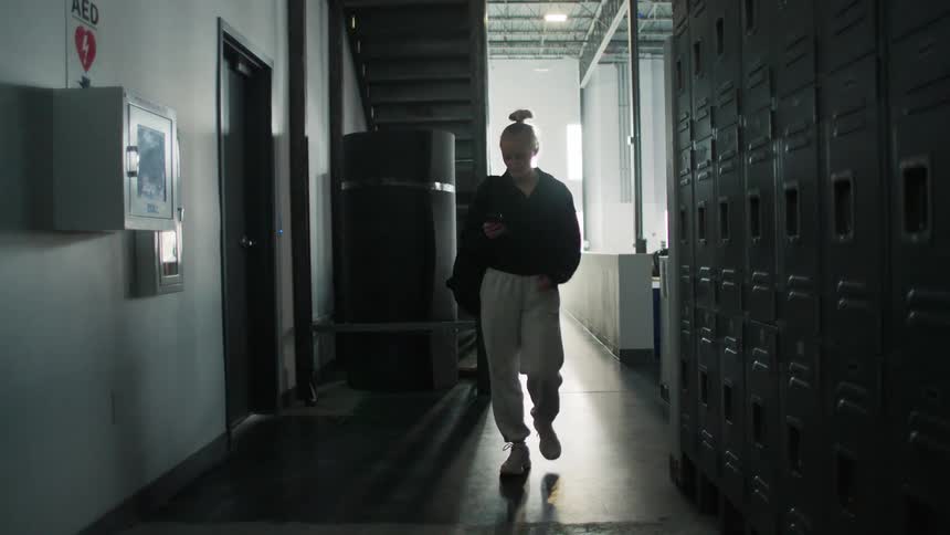 A young woman walks toward the camera down a dimly lit industrial corridor lined with metal lockers while focused on her smartphone screen
