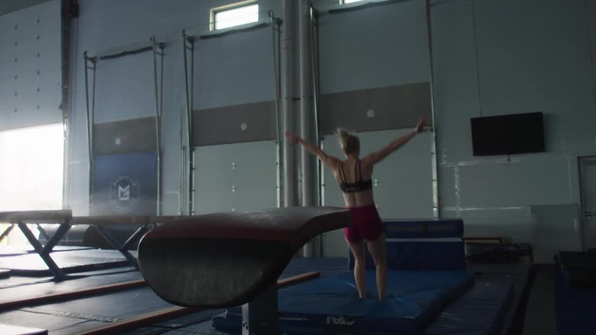 Inside a large gymnastics facility a female athlete executes a precise backflip dismount from a vaulting table sticking the landing on a thick blue mat