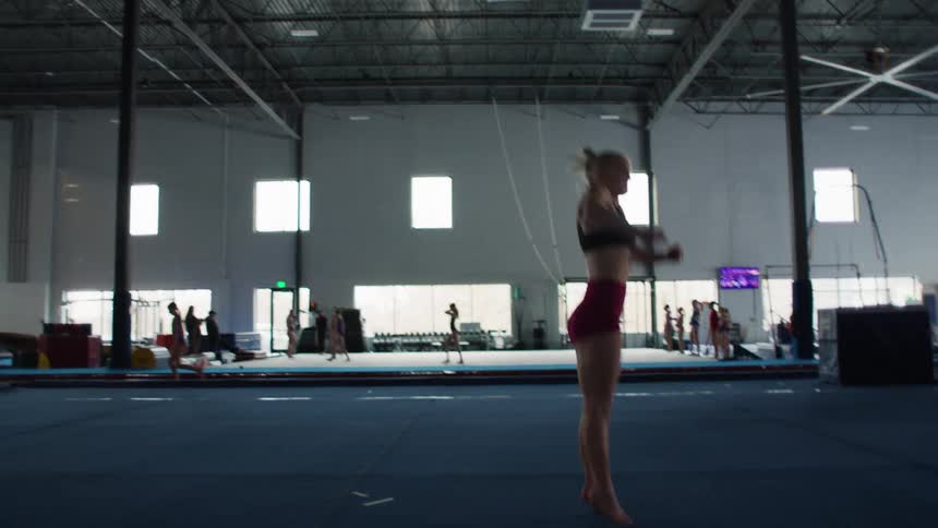 A female athlete wearing red shorts and a black sports bra runs and performs a high horizontal split jump and then another vertical split jump on a large blue mat in a vast brightly lit indoor gymnastics training facility