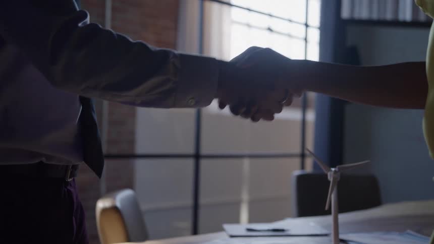 Two individuals in business attire are seen shaking hands over a conference table containing documents and a model wind turbine with their hands and upper bodies silhouetted against a bright large window in the background