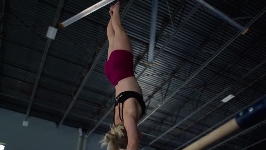 A female gymnast in a black sports bra and maroon shorts performs a routine on a single high gymnastics bar transitioning from a low swing into a handstand within the open space of a brightly lit training facility