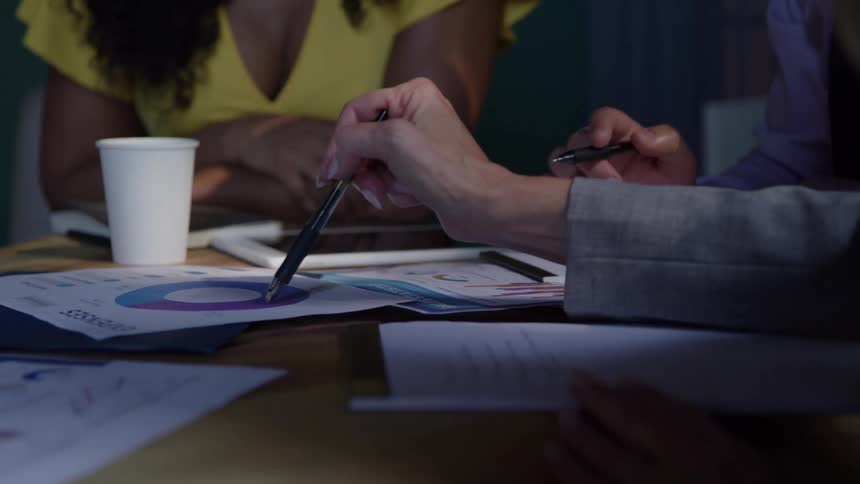 Female hands writing on a notepad and later pointing to a graph on a piece of paper while two other people take notes indoors