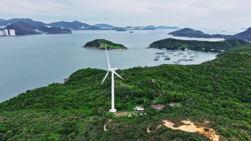An aerial wide shot shows a tall white wind turbine standing on a densely forested green hilltop overlooking a bay with various small islands floating structures and a dense cityscape backed by mountains visible across the water