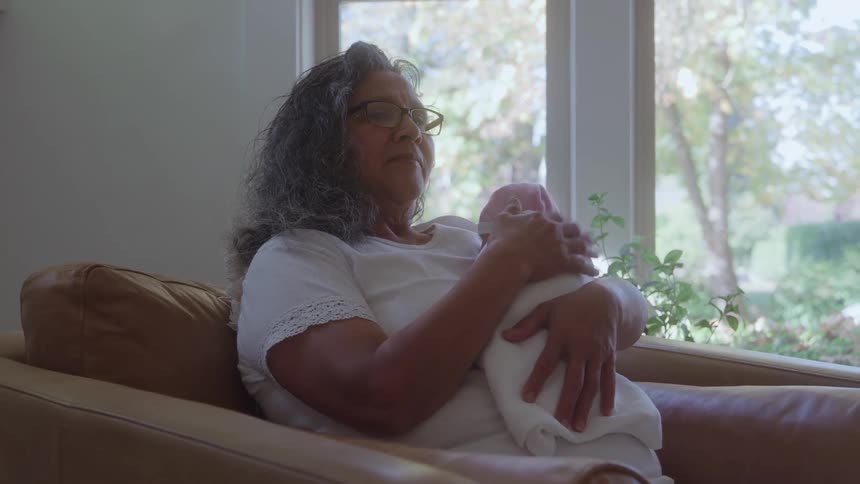 An older woman with gray curly hair and glasses a grandmother sits holding a swaddled infant near a sunlit window with a noticeable look of calm tenderness