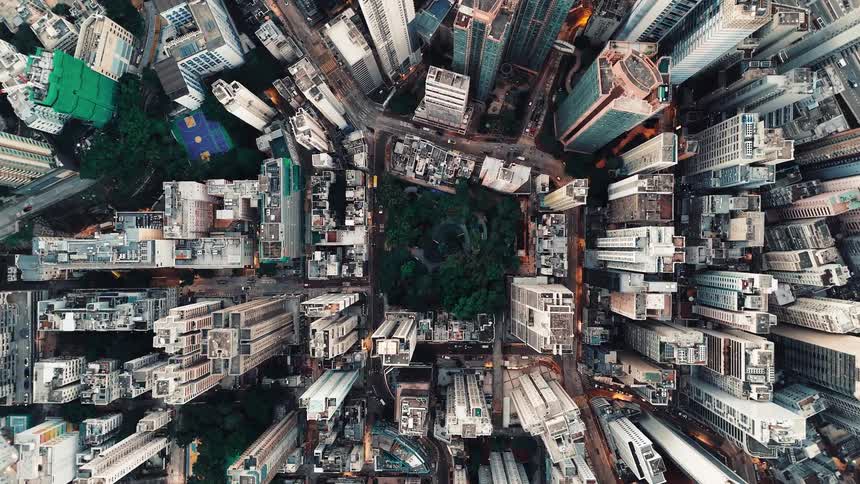 A dizzying topdown aerial shot reveals a dense metropolitan landscape where a small square of green trees is boxed in by towering skyscrapers and tight city streets