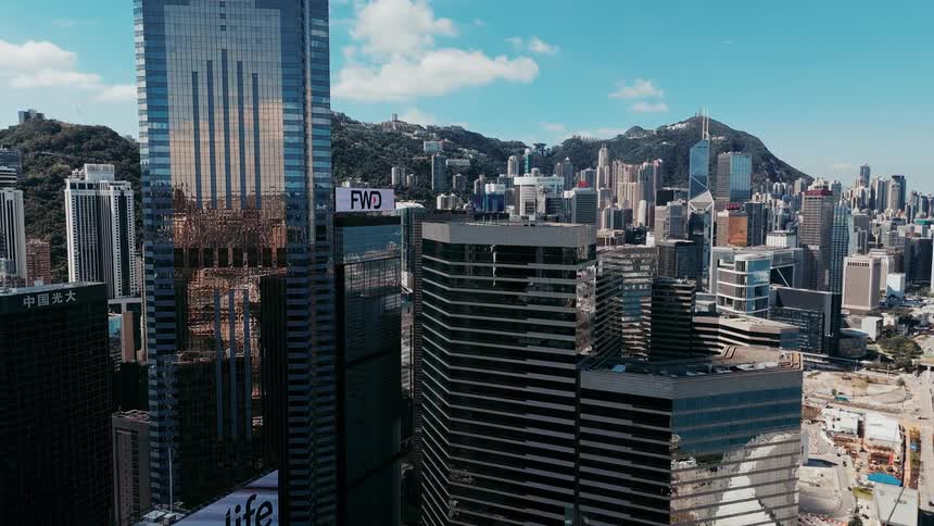 An aerial shot sweeps down past the towering reflective glass facades of modern skyscrapers showcasing a dense bustling city center built right against the base of steep forested hills under a bright blue sky with white clouds