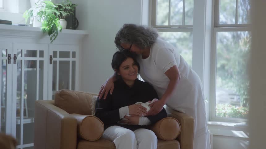 An older woman gently places her hand on a younger womans shoulder kissing the top of her head as she sits in a chair holding a newborn baby