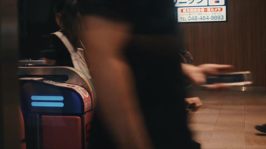A medium shot focuses on a row of colorful modern transit turnstiles as several people walk through the gates in an indoor station area
