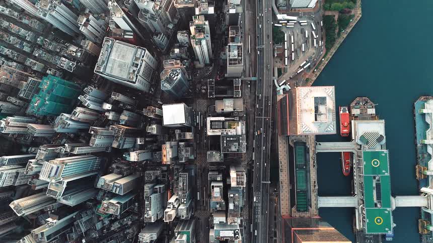 An extreme aerial overhead view pans over a dense urban city with highrise buildings separated by a road leading to a waterfront area with boat slips and docks