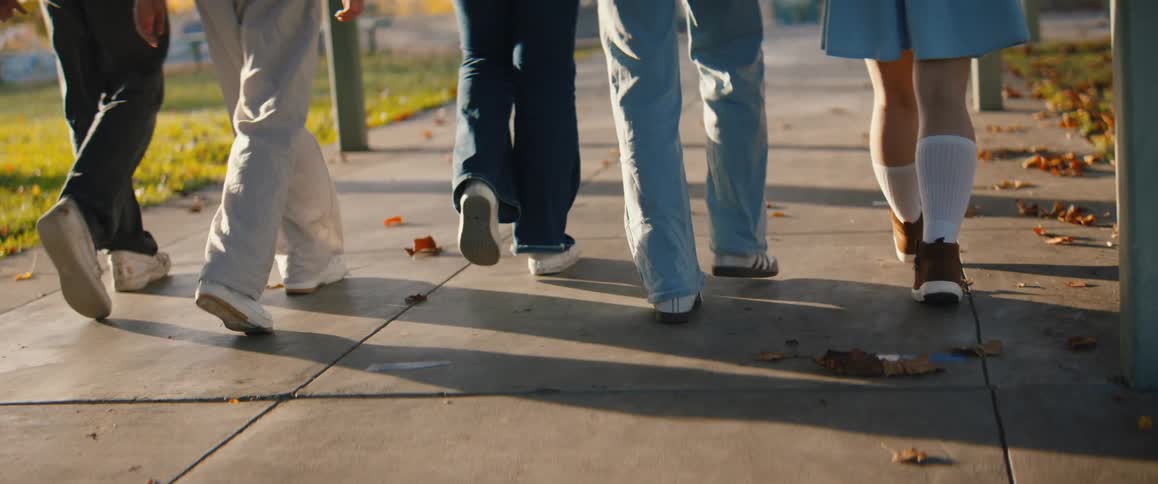This is a lowangle closeup shot focusing on the lower legs and feet of four individuals walking away from the camera on a paved sidewalkthey are dressed in casual pants sweatpants and jeans and varying footwear walking in dappled sunlight that ca