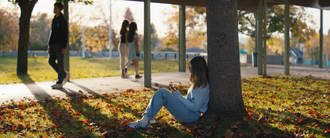 A young woman sits alone on the grass leaning against the base of a tree in a public outdoor area while other people walk by in the sunny background