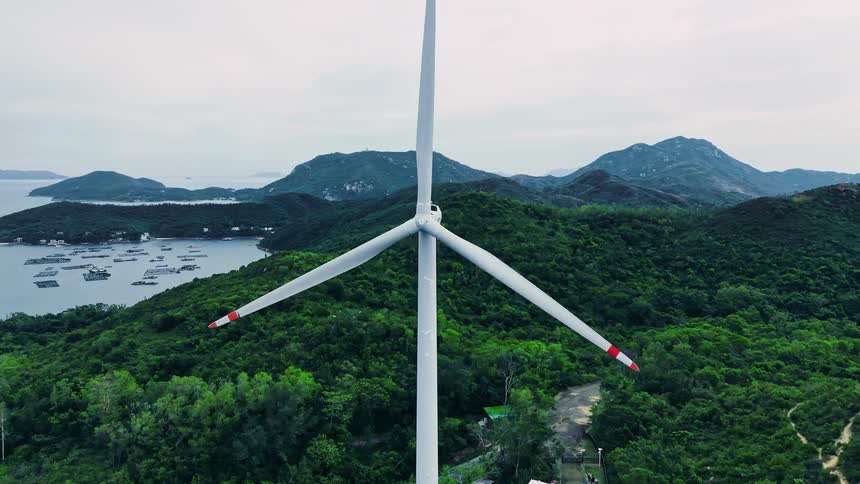 A tall white wind turbine with redtipped blades slowly rotating atop a densely forested green hill providing a sweeping view of the surrounding hilly coastline numerous small islands with a distant city visible on the horizon