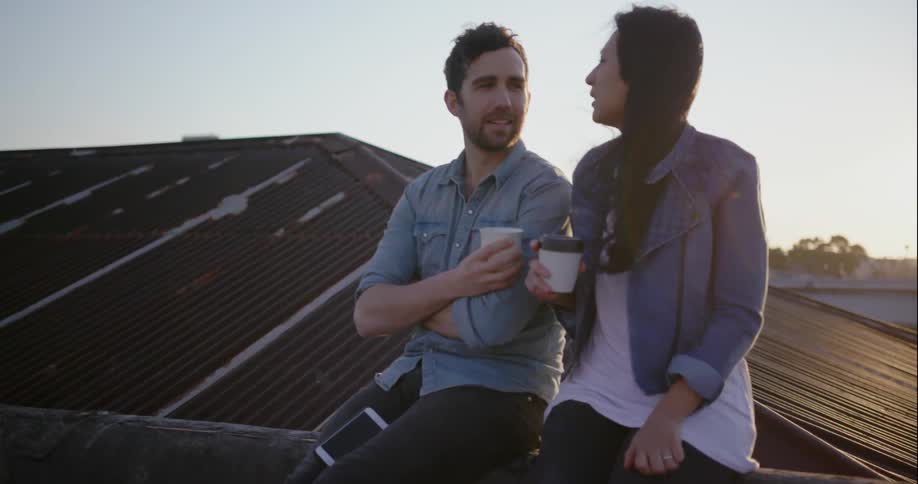 Two people sit on a roof drinking coffee