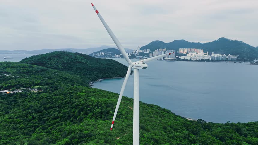 A massive white wind turbine steadily rotates its blades atop a lush green ridge overlooking a calm blue bay and a distant coastal city skyline