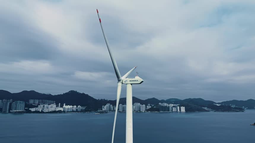 Wind turbine close up off the coast with mountains and waterfront buildings in the distance