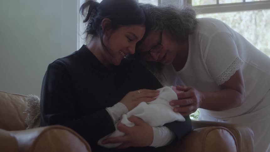 Woman seated holding a swaddled baby while another woman leans in close