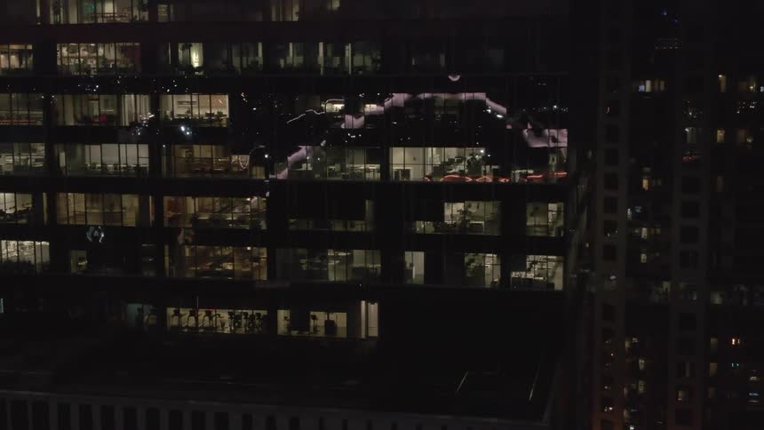 A large dark office building at night is seen with several floors brightly lit from within while a ghostly animated light projection of abstract shapes flickers and moves across its dark glass facade